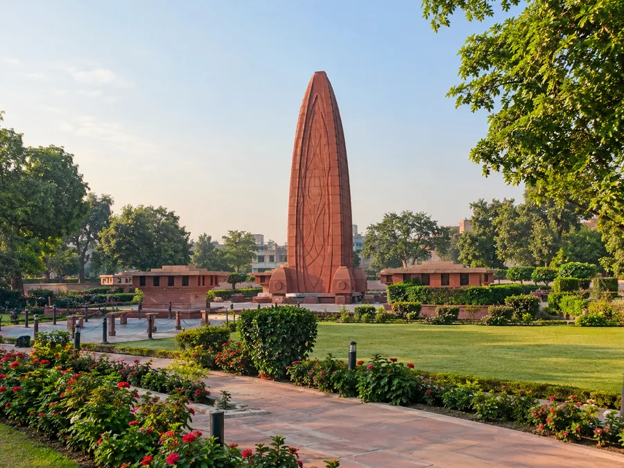 जलियांवाला बाग में लॉन और फूलों की क्यारियों के बीच लाल बलुआ पत्थर का ऊँचा, लौ के आकार का स्मारक